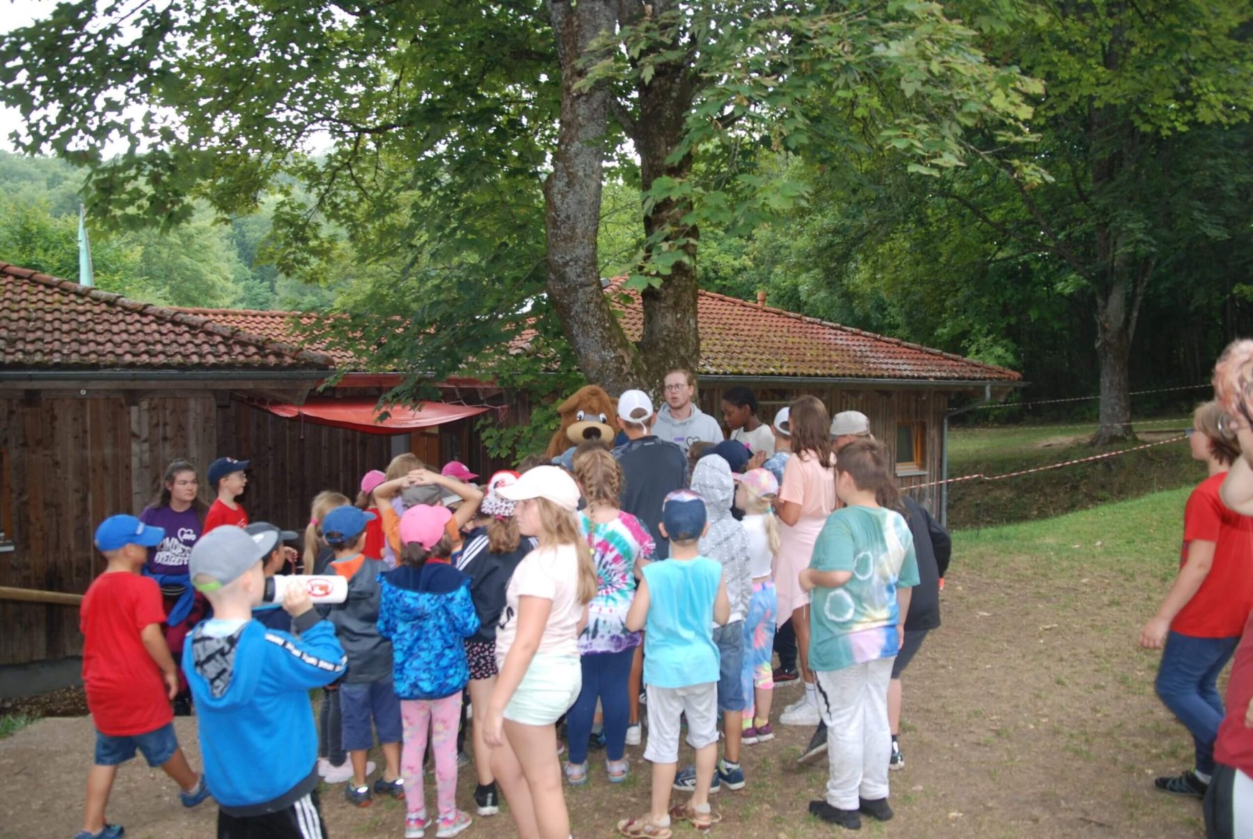 Kinder versammeln sich vor einer Holzhütte, um von einem Betreuer und dem Maskottchen Bär begrüßt zu werden.