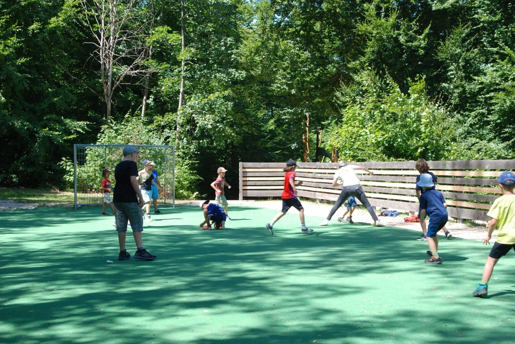 Kinder spielen mit einem Ball auf einem abgegrenzten Spielfeld unter schattigen Bäumen.