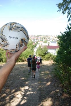 Eine Gruppe wandert über einen schmalen Pfad, während eine Hand im Vordergrund einen Fußball hält.