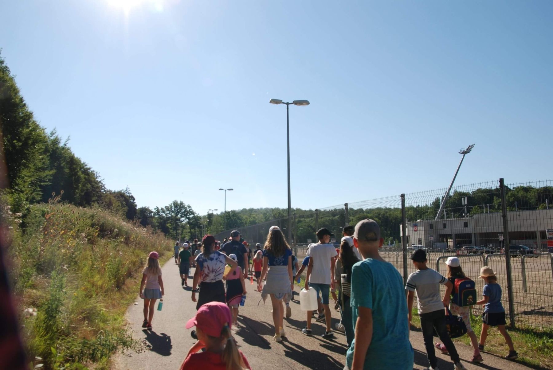 Kinder und Betreuer gehen entlang eines Weges vorbei an einem Sportplatz im Hintergrund.