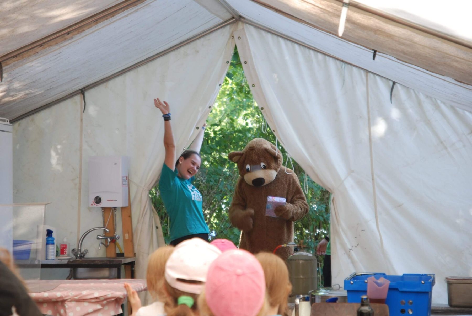 Eine Betreuerin und ein als Bär verkleideter Charakter begrüßen Kinder fröhlich in einem Zelt.